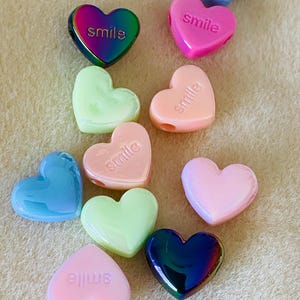 May include: A collection of heart-shaped beads in various colors including iridescent rainbow, pastel pink, light blue, and mint green. Several beads feature the word smile embossed on the surface. These small, smooth, glossy plastic components are arranged.