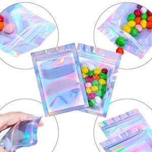 May include: A set of four iridescent holographic zippered pouches. The pouches are made of a clear plastic material with a holographic finish. The pouches are shown in different positions, including one with colorful candies inside.