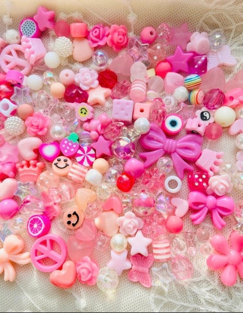 May include: A large assortment of pink and white beads, charms, and other decorative elements. The beads are made of various materials, including plastic, glass, and metal. Some of the charms include hearts, stars, flowers, and bows. The beads and charms are perfect for making jewelry, keychains, or other crafts.