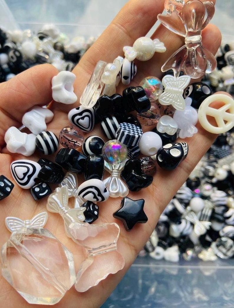 May include: A collection of black and white plastic beads in various shapes, including hearts, stars, bows, dice, and peace signs. The beads are in a hand, ready to be used for jewelry making or crafting.