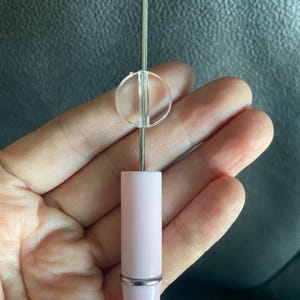 May include: A thin metal needle tool featuring a light pink handle and a small, clear, circular plastic disc attached to the shaft. The tool has a smooth, cylindrical design with a silver-toned metal band separating the handle from the needle.