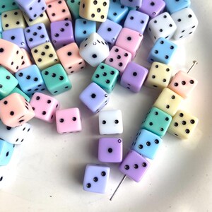 May include: A collection of colorful dice in pastel shades of pink, blue, purple, and yellow. Each die has black dots indicating the numbers. The dice are scattered on a white surface, creating a playful and visually appealing arrangement.