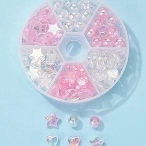 May include: A round plastic container with six compartments filled with iridescent pink, white, and clear beads, stars, and hearts. There are also loose beads, stars, and hearts on a light blue surface.