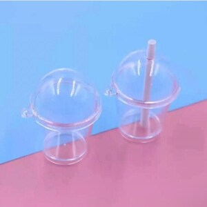 May include: Two clear plastic cups with domed lids and attached handles. One cup has a white straw. The cups are on a pink and blue background. The cups are designed for cold beverages.