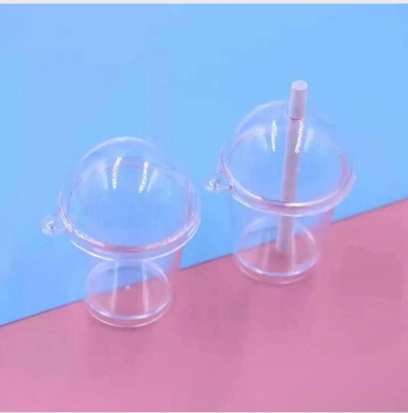 May include: Two clear plastic cups with domed lids and attached handles. One cup has a white straw. The cups are on a pink and blue background. The cups are designed for cold beverages.