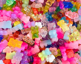 May include: A large collection of small, translucent gummy bear-shaped charms in a vibrant assortment of colors including pink, blue, yellow, green, orange, and purple. The charms feature a smooth, glossy finish and are scattered in a dense pile.