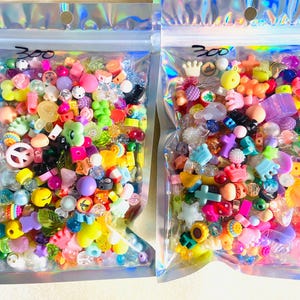 May include: Two clear plastic bags filled with colorful beads and charms. The beads are various shapes and sizes, including stars, flowers, and peace signs. The bags are sealed and have the number "300" written on them.