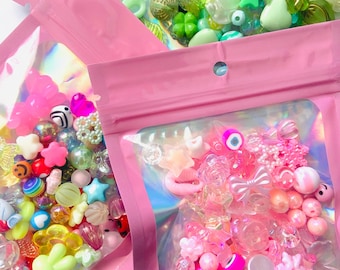 Puede incluir: Variadas cuentas de colores en bolsas de plástico transparente con cremalleras rosas. Las cuentas incluyen estrellas, flores, corazones y otras formas en varios colores como rosa, verde, amarillo y azul. Las bolsas están dispuestas sobre una superficie blanca.
