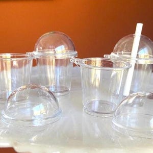 May include: A collection of clear plastic cups with dome lids, some with handles, and one with a white straw. The cups are arranged on a white, decorative surface, suggesting a beverage or dessert theme.