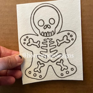 May include: A black and white skeleton gingerbread man design on a white square. The design includes a skull head, rib cage, and bone-shaped arms and legs. The text "@horrorpopdesigns" is visible.