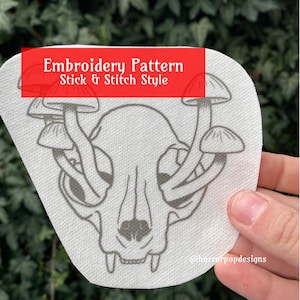 May include: A white embroidery pattern featuring a cat skull with mushrooms growing out of the top of the skull. The pattern is labeled "Embroidery Pattern Stick & Stitch Style" and "@horrorpopdesigns".