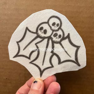 May include: A black and white illustration of holly leaves and berries, with the berries replaced by skulls. The design is on a white, textured paper. The text "@horrorpopdesigns" is visible on the image.