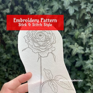 May include: A white embroidery pattern of a rose with an eye in the center. The pattern is labeled "Embroidery Pattern Stick & Stitch Style" and includes the social media handle "@horrorpopdesigns".