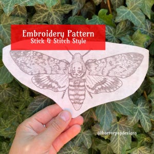 May include: Embroidery pattern of a hawk moth in a stick and stitch style. The pattern is printed on white paper and is held in a hand.