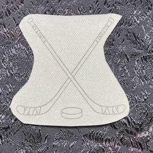 May include: A light gray patch featuring a hockey theme. Two crossed hockey sticks are depicted above a hockey puck. The patch has a unique shape and is set against a dark, patterned background.