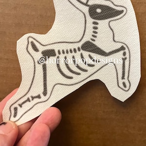 May include: A white, die-cut sticker featuring a black deer skeleton design. The deer is depicted in a running pose. The text "@horrorpopdesigns" is printed on the sticker.