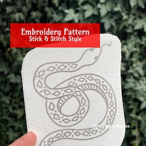 May include: A black and white embroidery pattern of a snake with a diamond pattern on its body. The pattern is on a white fabric background. The text "Embroidery Pattern Stick & Stitch Style" is at the top of the image.
