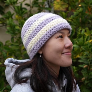May include: A purple and white striped crochet beanie hat. The hat is worn by a person with long dark hair.