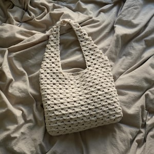 Op de afbeelding: Een witte gehaakte tote bag met een vierkant patroon. De tas ligt op een beige stoffen oppervlak.