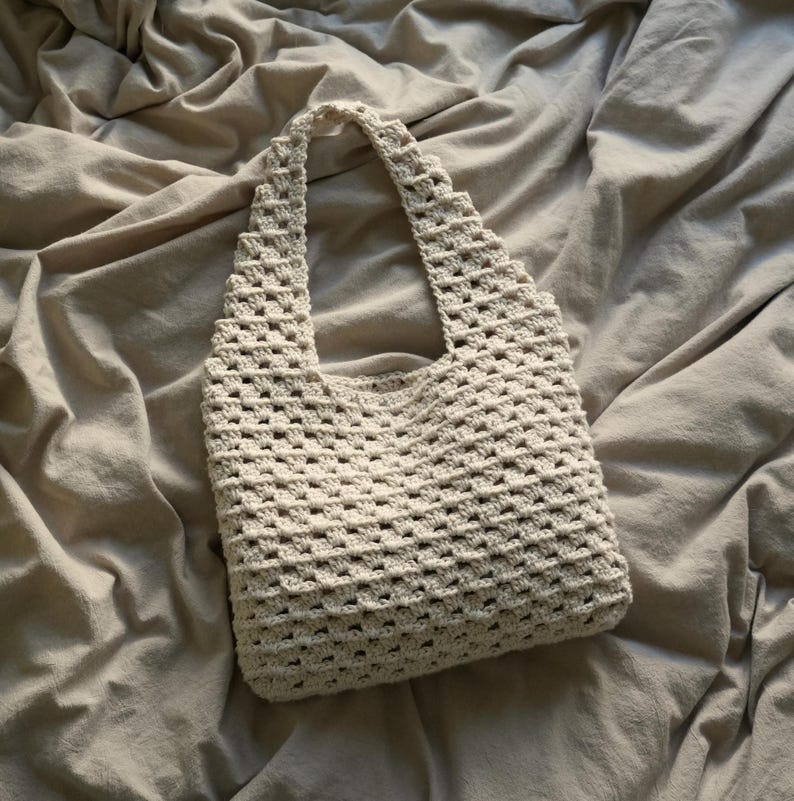 Op de afbeelding: Een witte gehaakte tote bag met een vierkant patroon. De tas ligt op een beige stoffen oppervlak.
