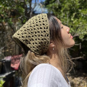 May include: A green crocheted bandana headband worn by a person with long brown hair. The headband is tied at the back of the head and the ends are hanging down.