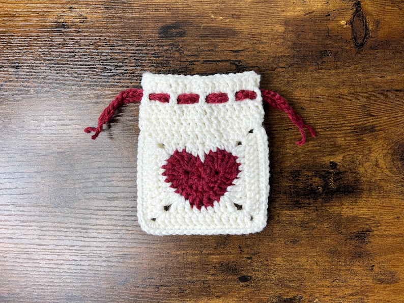 May include: A white crocheted drawstring bag with a red heart design. The bag is on a wooden surface.