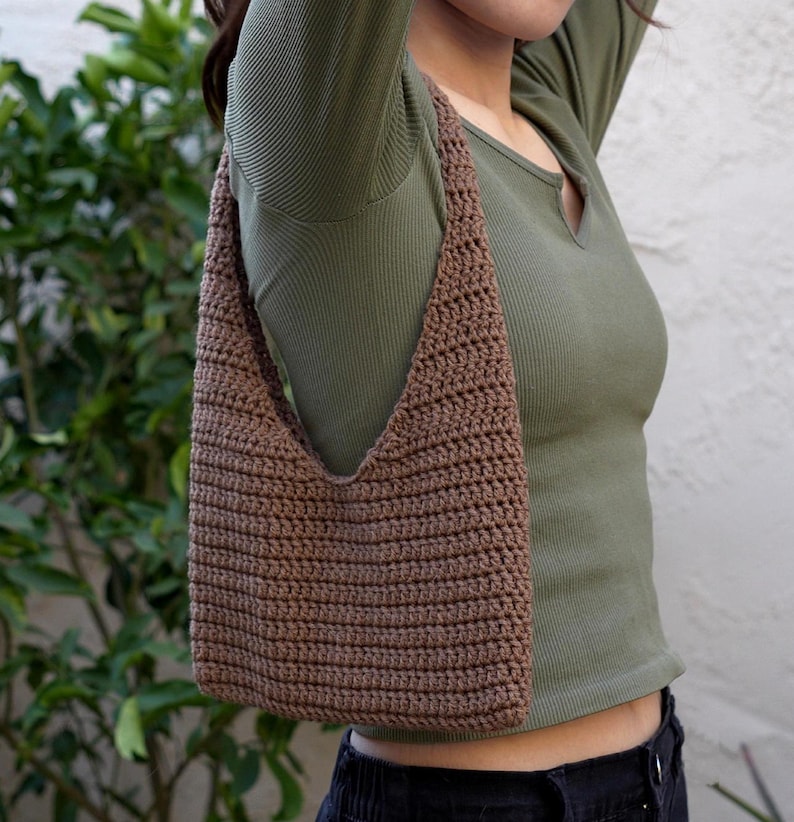 May include: A brown crocheted shoulder bag with a triangular shape. The bag is being worn by a person wearing a green long-sleeved shirt and black trousers.