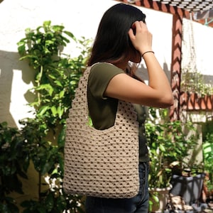 Op de afbeelding: Een beige gehaakte tote bag met een vierkant patroon. De tas wordt over de schouder gedragen door iemand die een groen shirt en jeans draagt.