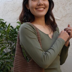 May include: A brown crocheted shoulder bag with a long strap. The bag is being worn by a person wearing a green long-sleeved shirt and black trousers.