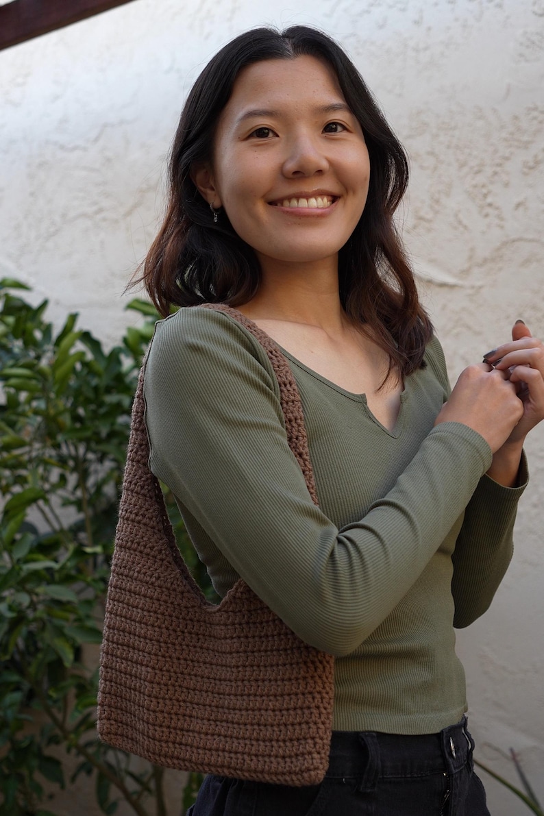 May include: A brown crocheted shoulder bag with a long strap. The bag is being worn by a person wearing a green long-sleeved shirt and black trousers.