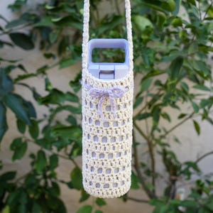 May include: A crocheted water bottle holder with a light brown color and a light purple ribbon. The holder is hanging from a light brown strap.
