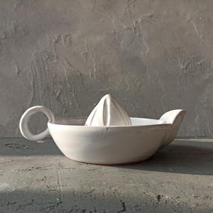 Op de afbeelding: Een witte keramische citruspers met een ingebouwde ruimer en een handvat. De pers heeft een komvorm met een tuit en is ontworpen voor gemakkelijke sapwinning. Het ontwerp is eenvoudig en functioneel.
