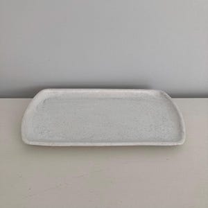 K&ouml;nnte beinhalten: Ein rechteckiges Keramiktablett mit gesprenkelter wei&szlig;er Oberfl&auml;che. Das Tablett verf&uuml;gt &uuml;ber leicht geschwungene, erh&ouml;hte R&auml;nder und eine flache, glatte Oberfl&auml;che. Das Design ist schlicht und funktional und weist eine helle, texturierte Optik auf.