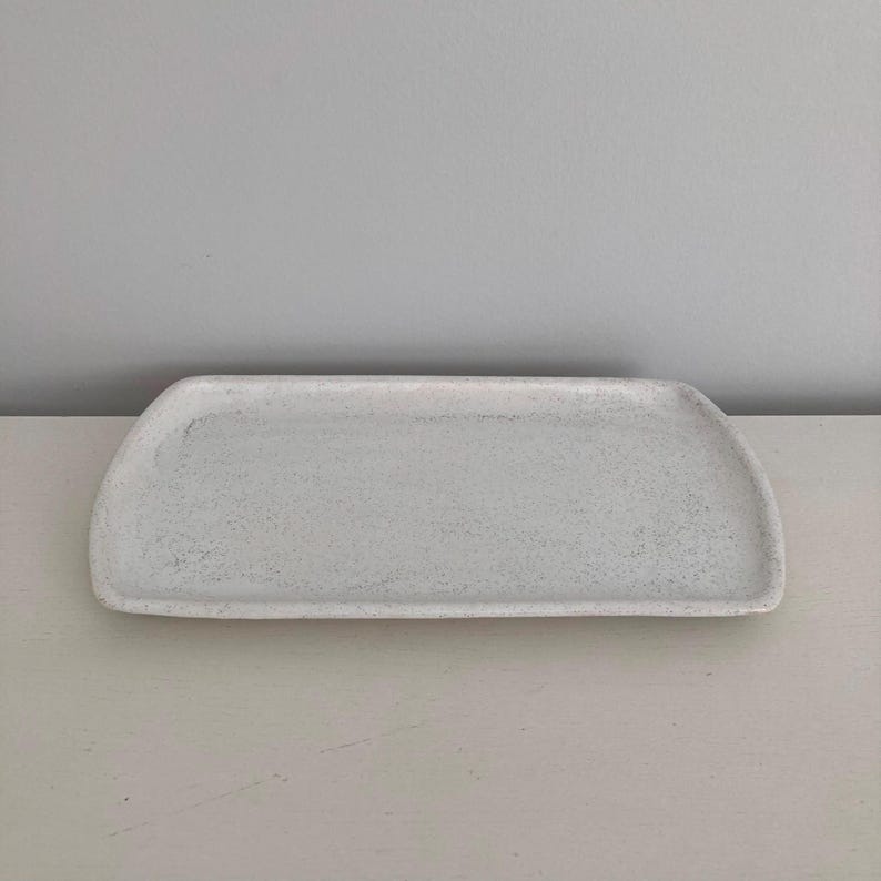 K&ouml;nnte beinhalten: Ein rechteckiges Keramiktablett mit gesprenkelter wei&szlig;er Oberfl&auml;che. Das Tablett verf&uuml;gt &uuml;ber leicht geschwungene, erh&ouml;hte R&auml;nder und eine flache, glatte Oberfl&auml;che. Das Design ist schlicht und funktional und weist eine helle, texturierte Optik auf.