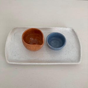 K&ouml;nnte beinhalten: Ein rechteckiges, wei&szlig; gesprenkeltes Keramiktablett mit zwei kleinen Schalen. Eine Schale ist ein abgerundetes orangefarbenes Gef&auml;&szlig; mit vertikalen Rippenstrukturen an der Au&szlig;enseite, w&auml;hrend die andere eine kleinere, glatte, blaue Schale ist.