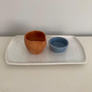 K&ouml;nnte beinhalten: Ein wei&szlig;es, gesprenkeltes, rechteckiges Keramiktablett mit einer terrakottafarbenen, gerippten Tasse und einer kleinen blauen Schale. Das Tablett hat abgerundete Ecken und eine flache Oberfl&auml;che, w&auml;hrend die Tasse vertikale Rillen aufweist.