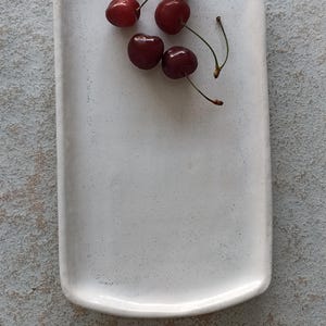 K&ouml;nnte beinhalten: Ein rechteckiges, cremewei&szlig;es Keramiktablett mit gesprenkelter Oberfl&auml;che und leicht erh&ouml;hten R&auml;ndern. F&uuml;nf frische, dunkelrote Kirschen mit gr&uuml;nen Stielen sind in einer kleinen Gruppe auf dem oberen Teil der glatten Fl&auml;che angeordnet.