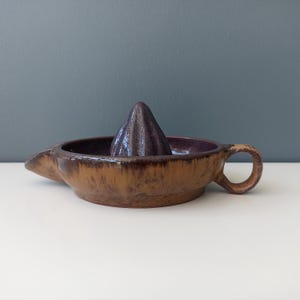 May include: A handmade ceramic citrus juicer with a dark brown and purple glaze. The juicer has a conical reamer in the center and a spout for pouring. The handle is round and the overall design is rustic.