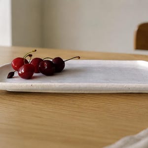 K&ouml;nnte beinhalten: Ein rechteckiges, cremewei&szlig;es Keramiktablett mit einem leicht unebenen, strukturierten Rand. Eine kleine Ansammlung von vier frischen, dunkelroten Kirschen mit gr&uuml;nen Stielen liegt auf der linken Seite der flachen, glatten Oberfl&auml;che.