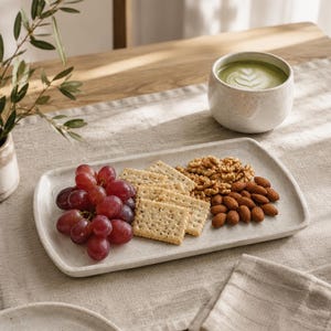Puede incluir: Una bandeja de servir rectangular de cerámica color blanco roto con motas contiene un racimo de uvas rojas, varias galletas rectangulares con semillas visibles, nueces y almendras enteras, junto a una taza de cerámica a juego.