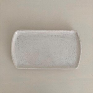K&ouml;nnte beinhalten: Ein rechteckiges Keramiktablett mit abgerundeten Ecken und einer gesprenkelten, cremewei&szlig;en Oberfl&auml;che. Die Oberfl&auml;che hat ein subtil texturiertes Aussehen mit kleinen dunklen Flecken, die &uuml;ber die helle Basis verteilt sind, was ein schlichtes Design ergibt.