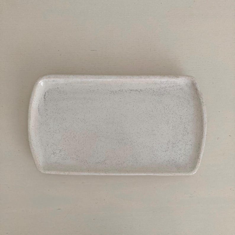 K&ouml;nnte beinhalten: Ein rechteckiges Keramiktablett mit abgerundeten Ecken und einer gesprenkelten, cremewei&szlig;en Oberfl&auml;che. Die Oberfl&auml;che hat ein subtil texturiertes Aussehen mit kleinen dunklen Flecken, die &uuml;ber die helle Basis verteilt sind, was ein schlichtes Design ergibt.