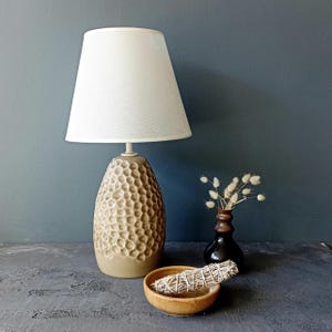 Könnte beinhalten: Eine beige Keramik-Tischlampe mit einem weißen Stofflampenschirm. Die Lampenbasis hat ein strukturiertes, wabenförmiges Design. Die Lampe steht auf einer grauen Oberfläche mit einer kleinen Schale mit getrocknetem Salbei und einer braunen Vase mit getrockneten Blumen.