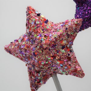 May include: A pink and red glitter star with a silver stick attached to the back. The star is made of a soft, textured material and is covered in glitter.
