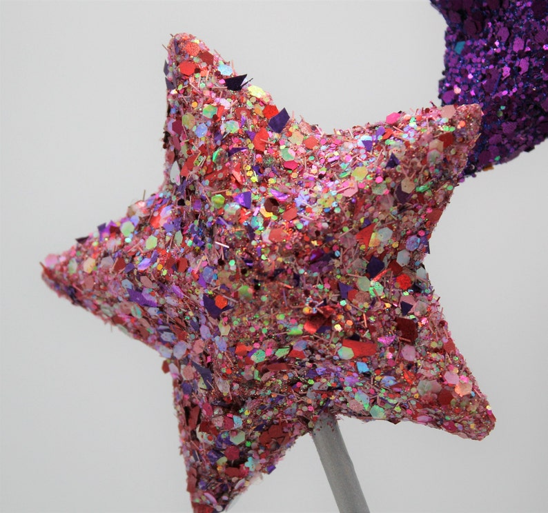 May include: A pink and red glitter star with a silver stick attached to the back. The star is made of a soft, textured material and is covered in glitter.