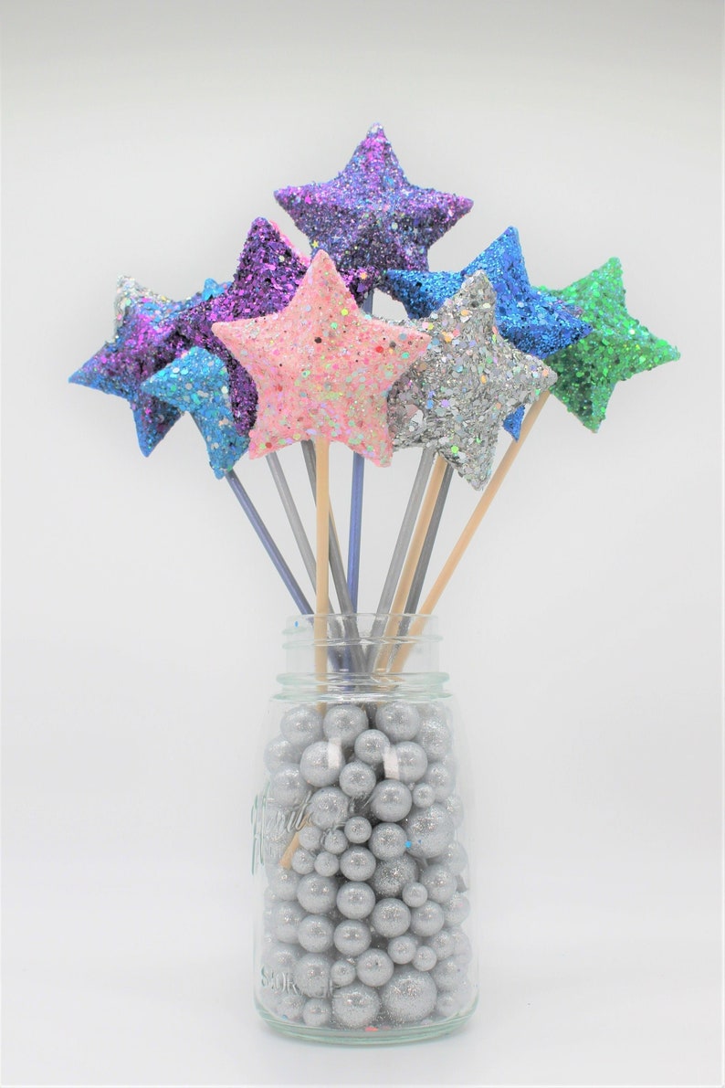May include: A glass jar filled with silver glitter balls, holding a bouquet of sparkly star-shaped decorations on wooden sticks. The stars are in various colors, including pink, blue, purple, green, and silver.