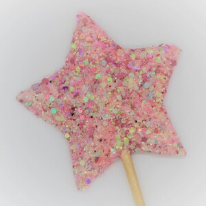 May include: A pink glitter star-shaped topper on a wooden stick. The star is covered in pink, white, and iridescent glitter.