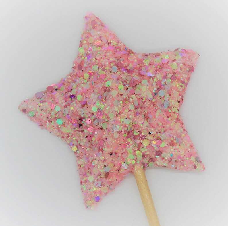 May include: A pink glitter star-shaped topper on a wooden stick. The star is covered in pink, white, and iridescent glitter.