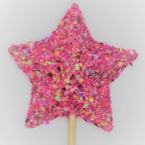 May include: A pink glitter star-shaped topper on a wooden stick. The star is covered in pink, gold, and iridescent glitter.