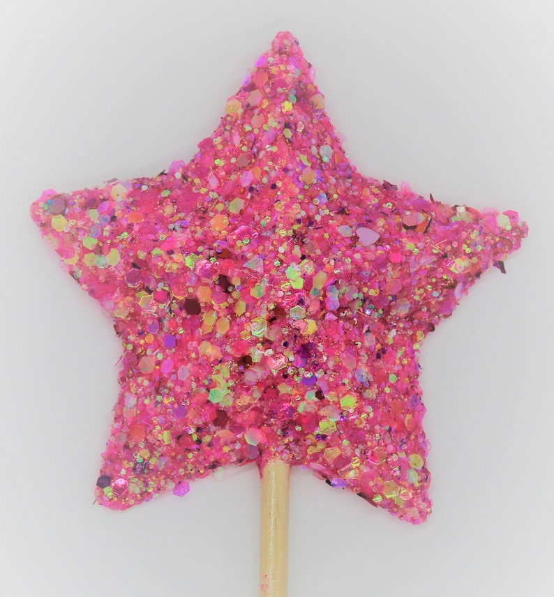 May include: A pink glitter star-shaped topper on a wooden stick. The star is covered in pink, gold, and iridescent glitter.